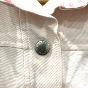 May include: Close-up of a white denim jacket with a pink and white striped collar. The jacket has a silver button with the word "Courr&egrave;ges" engraved on it.