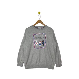 Puede incluir: Sudadera gris con un gr&aacute;fico de Hello Kitty y Gaspard et Lisa. El gr&aacute;fico est&aacute; en un marco rojo y azul con rayas blancas. El texto "Gaspard et Lisa Hello Kitty" est&aacute; impreso encima del gr&aacute;fico.