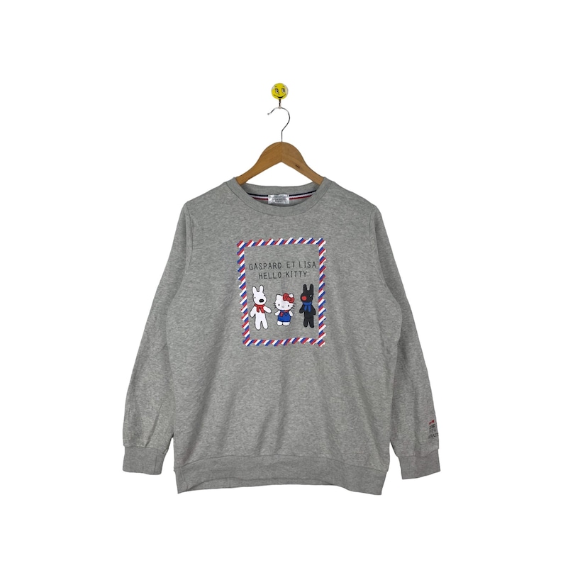 Puede incluir: Sudadera gris con un gr&aacute;fico de Hello Kitty y Gaspard et Lisa. El gr&aacute;fico est&aacute; en un marco rojo y azul con rayas blancas. El texto "Gaspard et Lisa Hello Kitty" est&aacute; impreso encima del gr&aacute;fico.