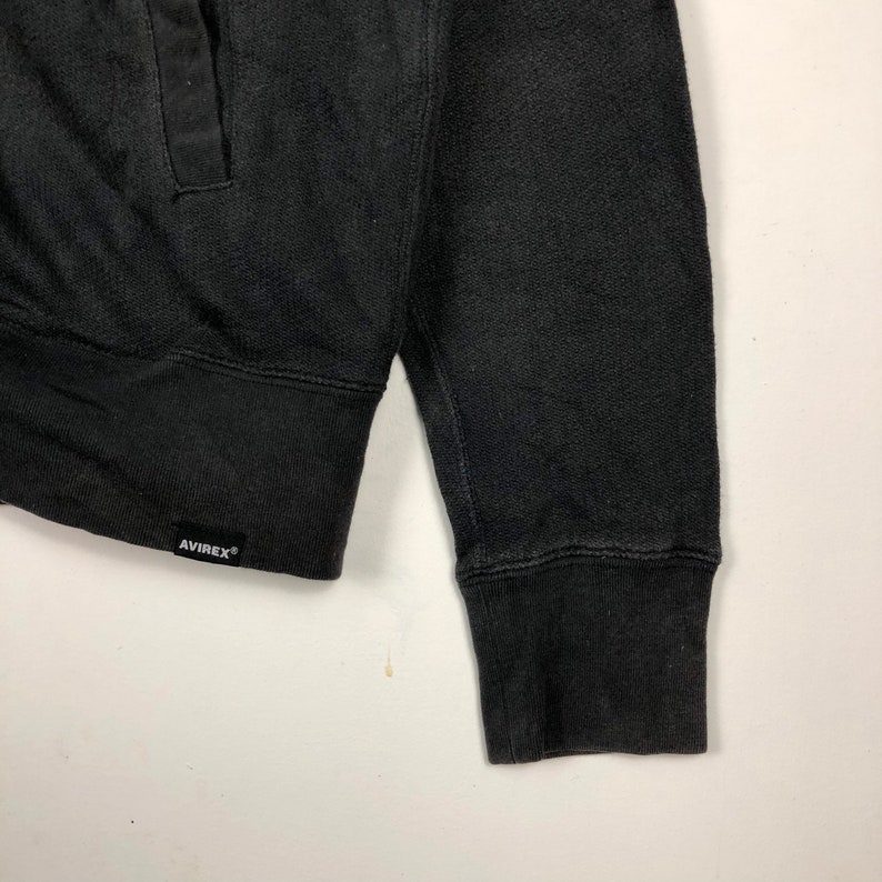 May include: Black long-sleeve sweatshirt with a ribbed cuff and a black label with the text "AVIREX" on the bottom left side.