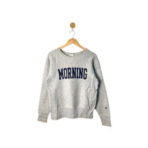 Puede incluir: Sudadera Champion gris claro con la palabra "MORNING" en letras mayúsculas azul marino en el pecho. La sudadera tiene cuello redondo, mangas largas y un bolsillo delantero. Un pequeño logotipo de Champion es visible en la manga izquierda.