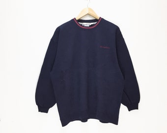 May include: Navy blue sweatshirt with a crew neck and long sleeves. The collar has a red and white trim. The text "et originals" is embroidered in red on the chest. The sweatshirt is hanging on a wooden hanger.