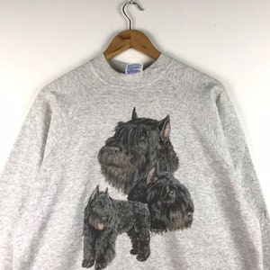 May include: A gray sweatshirt with a black and white graphic print of three Bouvier des Flandres dogs. The dogs are in different poses, with one dog's head facing the viewer, another dog's head facing to the right, and the third dog standing with its head facing to the left.