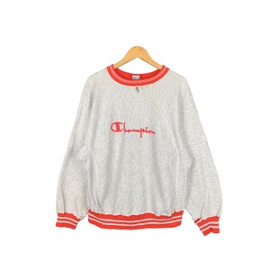 Puede incluir: Una sudadera gris Champion con ribetes rojos alrededor del cuello y los puños. El logotipo de Champion está bordado en rojo en la parte delantera.
