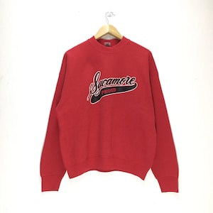 May include: A red sweatshirt featuring the word "Sycamore" in a script font and the word "Proud" underneath. The text is in black with a white outline and a black angled background. The sweatshirt is hanging on a wooden hanger.