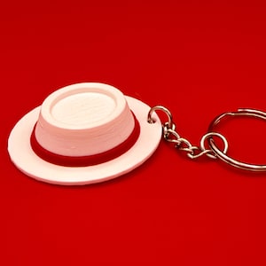 May include: A white miniature hat keychain featuring a flat brim and a rounded crown. A thin, vibrant red band encircles the base of the crown. The hat is attached to a silver-toned metal chain and a circular split ring for keys.