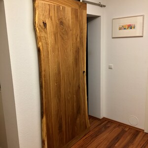 Puede incluir: Una gran puerta de granero de madera natural con borde vivo, montada sobre una gu&iacute;a corredera de acero inoxidable. La puerta es de color marr&oacute;n claro c&aacute;lido con vetas y nudos de madera visibles. La puerta est&aacute; parcialmente abierta, revelando un interior oscuro.