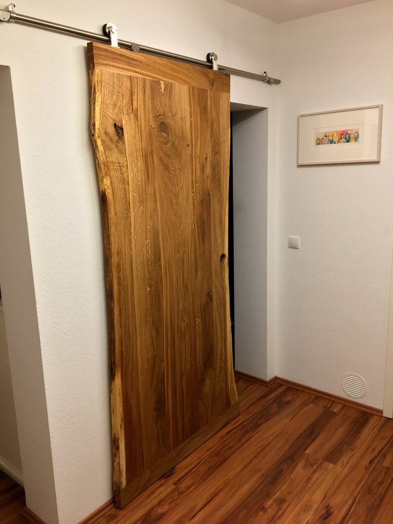 Puede incluir: Una gran puerta de granero de madera natural con borde vivo, montada sobre una gu&iacute;a corredera de acero inoxidable. La puerta es de color marr&oacute;n claro c&aacute;lido con vetas y nudos de madera visibles. La puerta est&aacute; parcialmente abierta, revelando un interior oscuro.