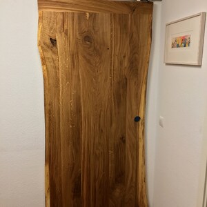 Puede incluir: Una puerta corredera de granero de madera natural con borde en bruto. La puerta est&aacute; montada sobre una gu&iacute;a de acero inoxidable y tiene un tirador azul oscuro. La veta de la madera es visible y la puerta es de color marr&oacute;n c&aacute;lido.