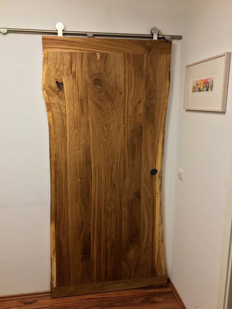 Puede incluir: Una puerta corredera de granero de madera natural con borde en bruto. La puerta est&aacute; montada sobre una gu&iacute;a de acero inoxidable y tiene un tirador azul oscuro. La veta de la madera es visible y la puerta es de color marr&oacute;n c&aacute;lido.