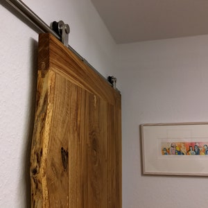 Puede incluir: Una puerta de granero de madera r&uacute;stica con borde natural, montada sobre una gu&iacute;a corredera de acero inoxidable. La puerta es de color marr&oacute;n c&aacute;lido y tiene vetas de madera visibles. Una obra de arte enmarcada cuelga de la pared blanca.