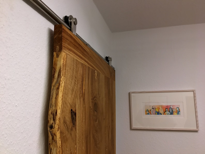 Puede incluir: Una puerta de granero de madera r&uacute;stica con borde natural, montada sobre una gu&iacute;a corredera de acero inoxidable. La puerta es de color marr&oacute;n c&aacute;lido y tiene vetas de madera visibles. Una obra de arte enmarcada cuelga de la pared blanca.