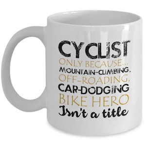 May include: White ceramic coffee mug with a black and gold design. The text on the mug reads "CYCLIST ONLY BECAUSE... MOUNTAIN-CLIMBING, OFF-ROADING, CAR-DODGING BIKE HERO Isn't a title"