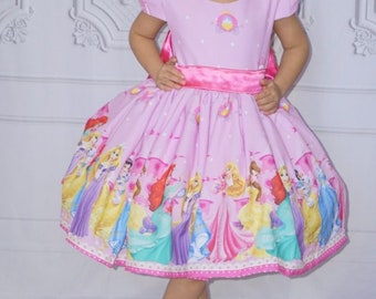 Puede incluir: Vestido de princesa rosa con una falda amplia con un estampado colorido de varias princesas. El vestido tiene mangas cortas, una faja rosa y un lazo a juego. Zapatos blancos completan el conjunto.