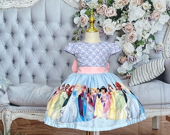 May include: A light blue dress with a princess-themed design. The bodice has a diamond pattern, a pink sash, and short sleeves. The skirt features various princess characters. The text "Baby Eden Boutique" is visible.
