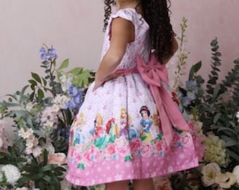 May include: A young girl wearing a pink and white dress with a princess theme. The dress features images of Disney princesses, including Cinderella, Snow White, and Ariel. The girl is standing in front of a floral backdrop.