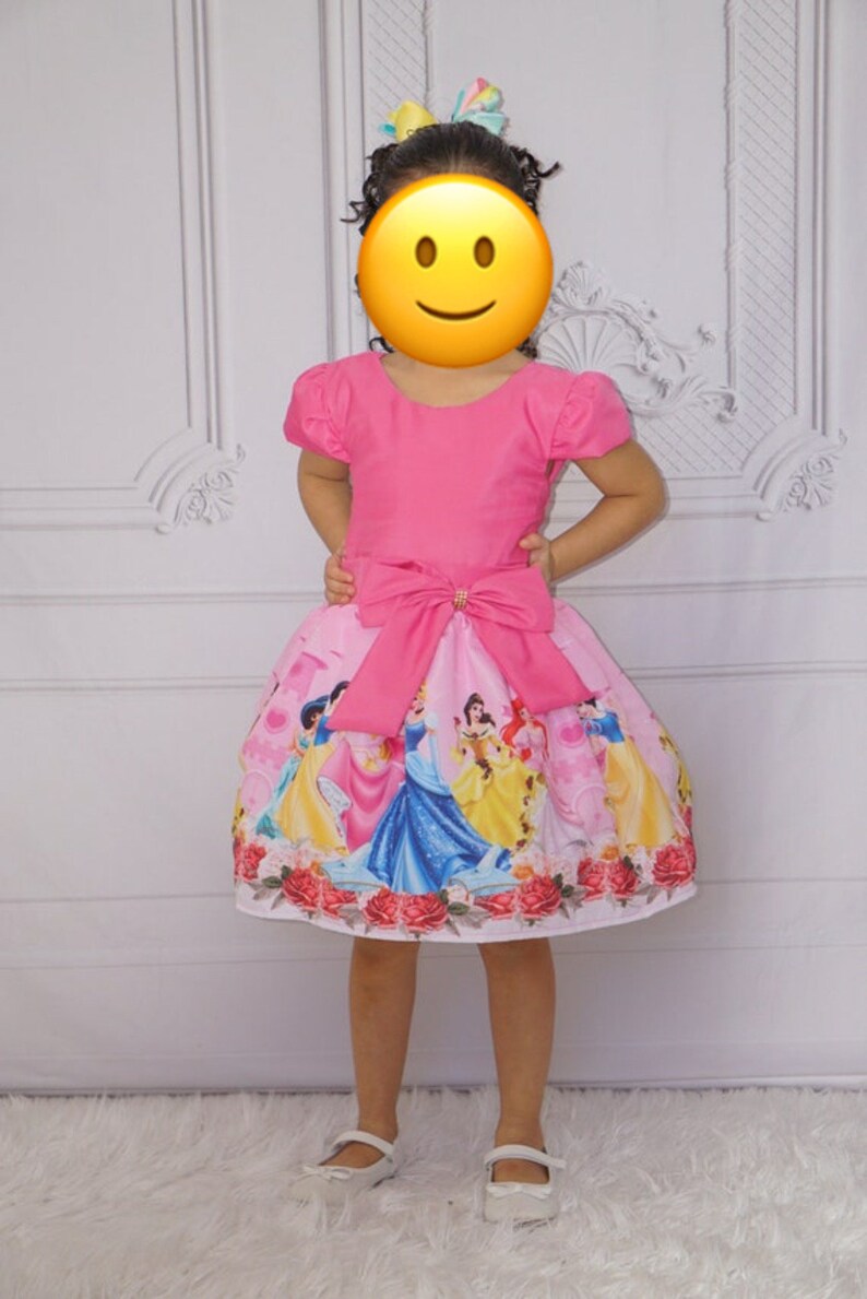 May include: A pink dress with short sleeves and a pink bow at the waist. The skirt of the dress has a print of Disney princesses.