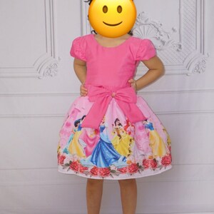 May include: A pink dress with short sleeves and a pink bow at the waist. The skirt of the dress has a print of Disney princesses.