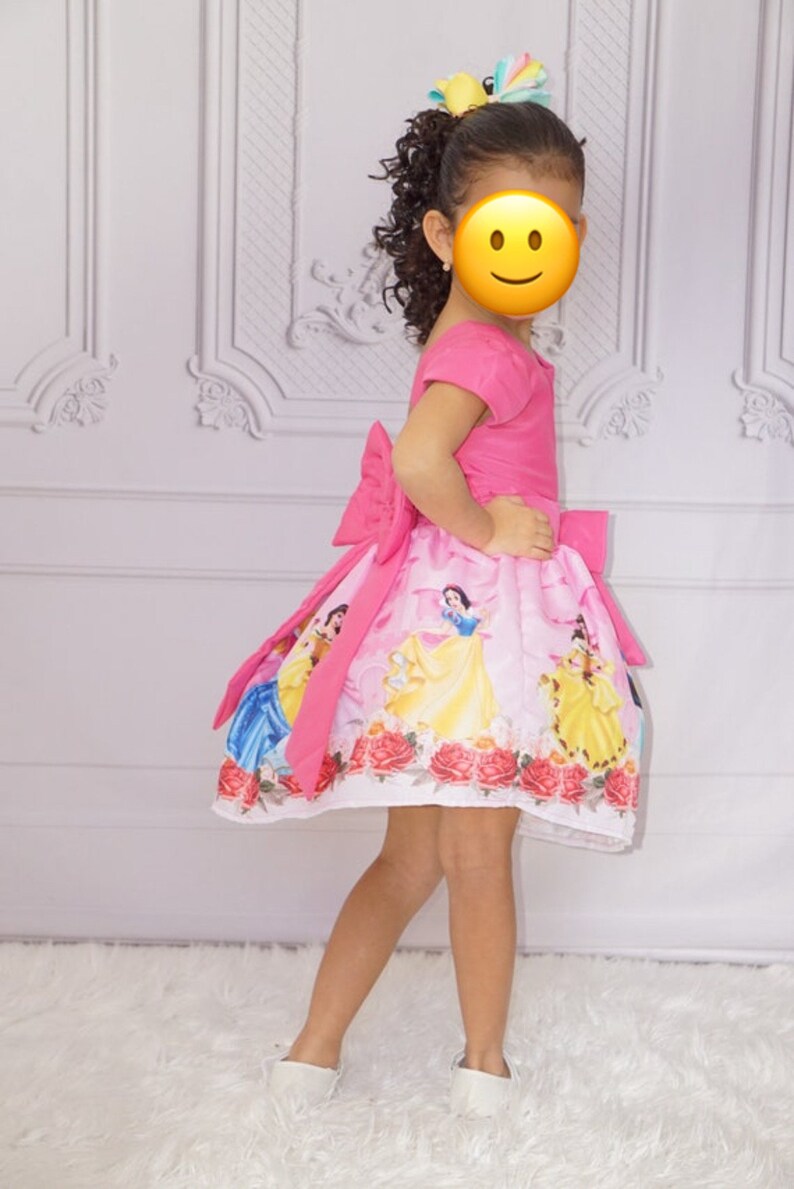 May include: A young girl wearing a pink dress with a white skirt featuring a print of Disney princesses. The dress has a large pink bow on the back. The girl is wearing white shoes and has her hair pulled back with a colorful bow.
