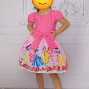 May include: A pink princess dress with a large pink bow at the waist. The skirt features a print of Disney princesses, including Cinderella, Snow White, and Belle. The dress is perfect for a little girl's birthday party or special occasion.
