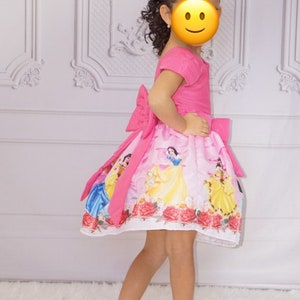 May include: A young girl wearing a pink dress with a white skirt featuring a print of Disney princesses. The dress has a large pink bow on the back. The girl is wearing white shoes and has her hair pulled back with a colorful bow.