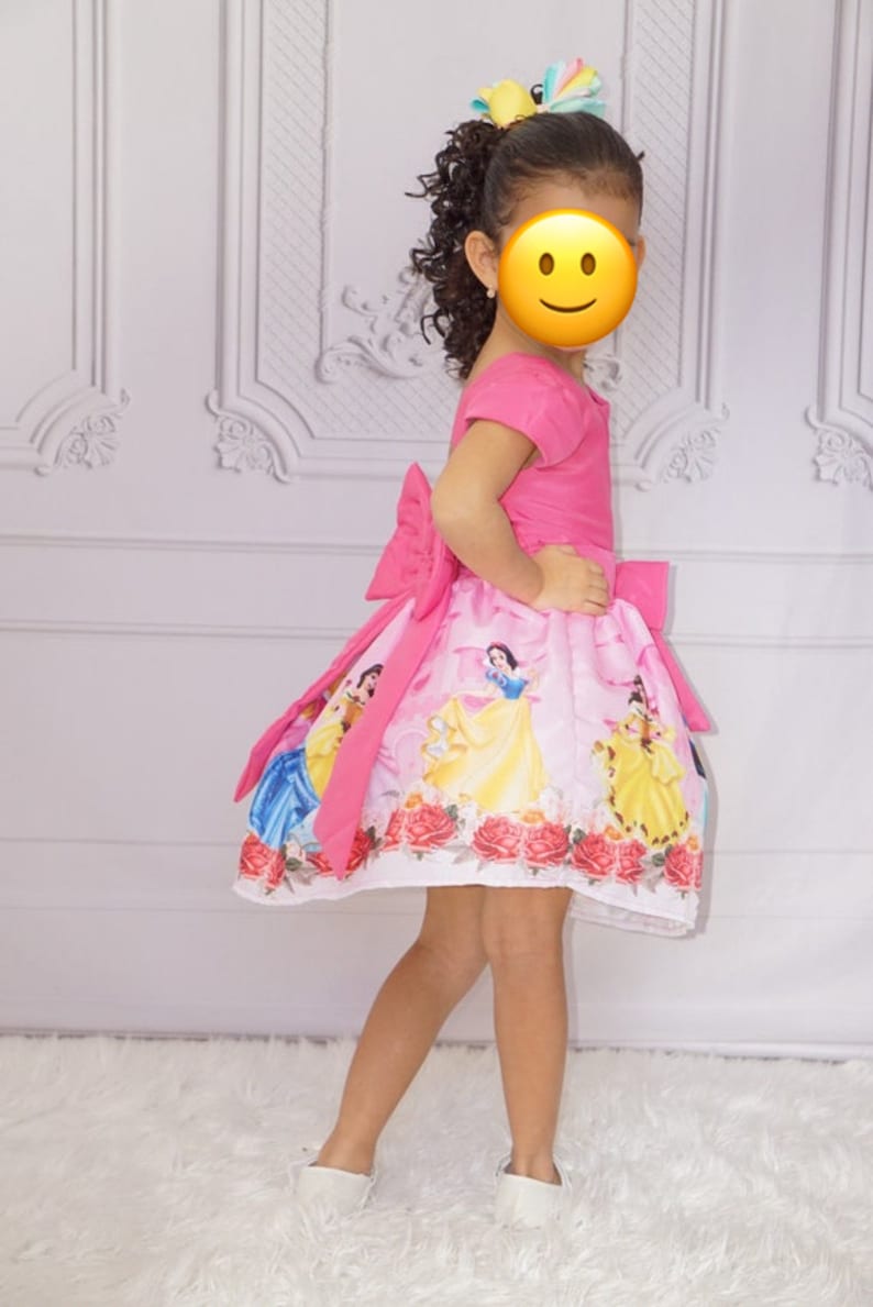 May include: A young girl wearing a pink dress with a white skirt featuring a print of Disney princesses. The dress has a large pink bow on the back. The girl is wearing white shoes and has her hair pulled back with a colorful bow.