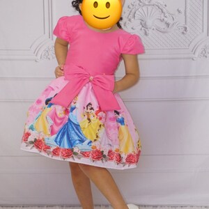 May include: A pink dress with short sleeves and a large pink bow at the waist. The skirt of the dress features a print of Disney princesses, including Cinderella, Snow White, Belle, and Ariel. The dress is worn by a young girl with curly hair and a colorful bow in her hair.