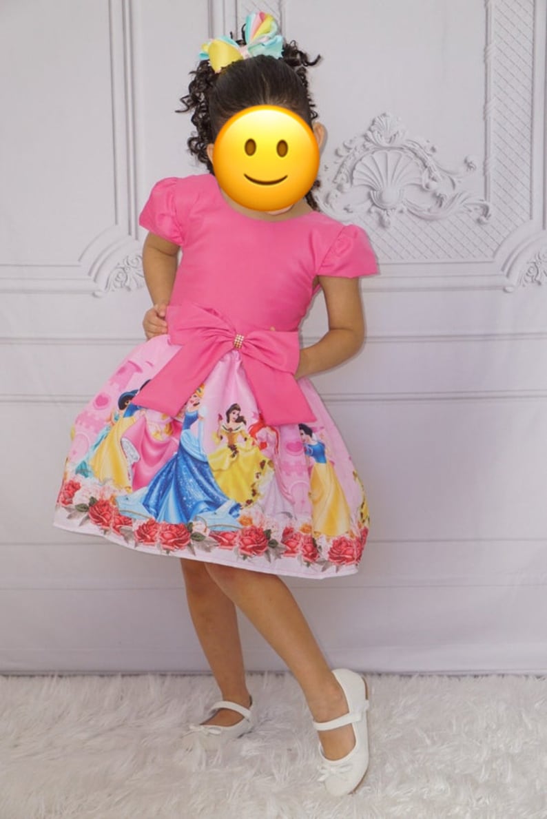 May include: A pink dress with short sleeves and a large pink bow at the waist. The skirt of the dress features a print of Disney princesses, including Cinderella, Snow White, Belle, and Ariel. The dress is worn by a young girl with curly hair and a colorful bow in her hair.