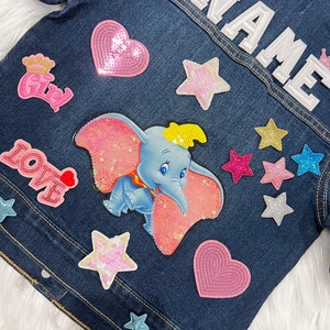 以下が含まれることがあります： 様々なカラフルなパッチが付いた青いデニムジャケット。ピンクのハート、ピンクの象、王冠、星、そして「LOVE」の文字。ジャケットの背面には「NAME」の文字が縫い付けられています。