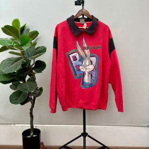 Op de afbeelding: Rood sweatshirt met zwarte bies en een Bugs Bunny-afbeelding. De afbeelding toont Bugs Bunny in een blauwe en groene "B" met de tekst "Bugs Bunny" erboven.