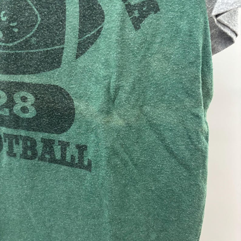 May include: A green t-shirt with heathered texture featuring a black graphic print of a football, the number 28, and the word FOOTBALL. The shirt has contrasting light gray sleeves and a crew neckline, showcasing a casual athletic design.