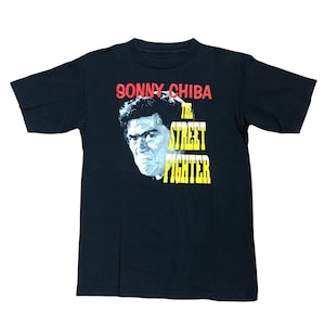 May include: Black t-shirt featuring a portrait of Sonny Chiba with the text "SONNY CHIBA" in red above the image and "THE STREET FIGHTER" in yellow below. The shirt has short sleeves and a crew neck.