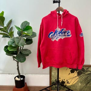 May include: A red hoodie with a front pocket and a drawstring hood. The front features the text "Joker WORLDWIDE" in blue and silver lettering. A smaller logo is on the sleeve. The hoodie is hanging on a black stand.