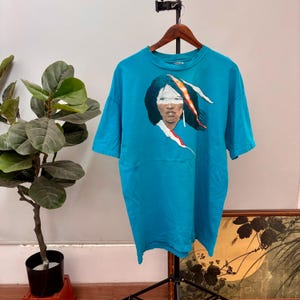 Peut inclure: Un t-shirt turquoise avec un graphisme d'une personne amérindienne aux longs cheveux noirs, au maquillage blanc et à une plume rouge et orange. Le t-shirt est suspendu à un cintre en bois.