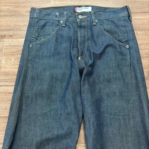 May include: Blue denim jeans with a relaxed fit. The jeans feature a classic five-pocket design, a button and zip closure, and a red tag that reads "Engineered Jeans Relaxed". The jeans are made of a durable denim fabric.