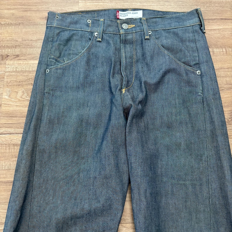 May include: Blue denim jeans with a relaxed fit. The jeans feature a classic five-pocket design, a button and zip closure, and a red tag that reads "Engineered Jeans Relaxed". The jeans are made of a durable denim fabric.