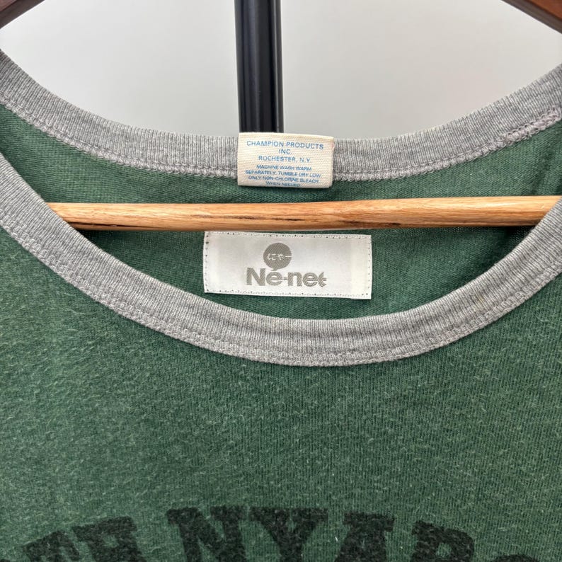 May include: A green ringer t-shirt with a grey ribbed crew neckline. The interior features two white labels: one reads Champion Products Inc. Rochester, N.Y. with care instructions, and the other displays the brand name N&eacute;-net with Japanese text.