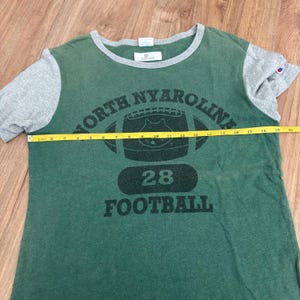 May include: A green and gray short-sleeve t-shirt with a graphic of a football and the text NORTH NYAROLINA 28 FOOTBALL. A yellow measuring tape is placed across the chest, showing a width of approximately 20 inches from edge to edge.