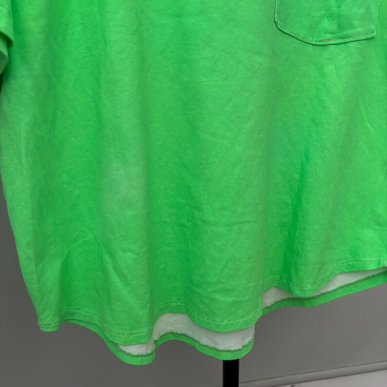 Puede incluir: Camiseta verde brillante con un bolsillo en la parte superior izquierda. La camiseta tiene un dise&ntilde;o en capas con una capa inferior blanca visible en el dobladillo. La tela tiene una apariencia ligeramente descolorida.