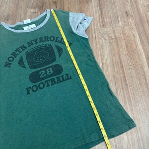 May include: A green t-shirt with gray sleeves and a gray collar, featuring a black graphic of a football with a cat face and the text NORTH NYAROLINA 28 FOOTBALL. A yellow measuring tape shows the shirt length is approximately 27 inches.