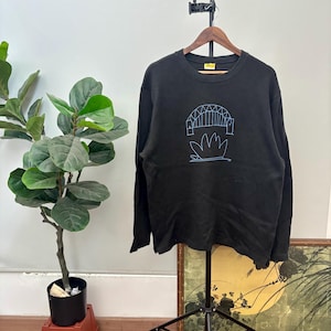 Könnte beinhalten: Schwarzes Langarm-Sweatshirt mit einer blauen Grafik einer Brücke und einer Pflanze. Das Sweatshirt hängt an einem Holzbügel. Im Hintergrund befinden sich eine Pflanze und ein gerahmtes Bild.