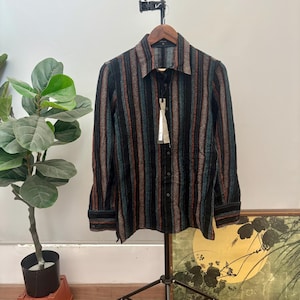 Pode incluir: Uma camisa de manga comprida com botões, apresentando um padrão de riscas verticais em tons de preto, azul-petróleo e castanho-ferrugem. A camisa tem um colarinho padrão, uma carcela de botões frontal e punhos abotoados num cabide.