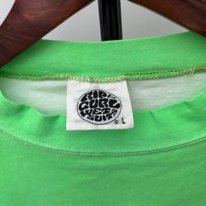 Puede incluir: Primer plano de una camiseta verde brillante con una etiqueta blanca. La etiqueta presenta un logotipo circular en blanco y negro con el texto "Rip Curl Wet Suits" y la talla "L". La camiseta est&aacute; colgada en una percha de madera.