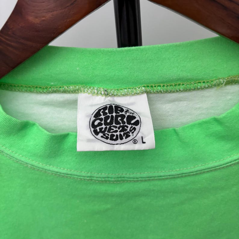 Puede incluir: Primer plano de una camiseta verde brillante con una etiqueta blanca. La etiqueta presenta un logotipo circular en blanco y negro con el texto "Rip Curl Wet Suits" y la talla "L". La camiseta est&aacute; colgada en una percha de madera.