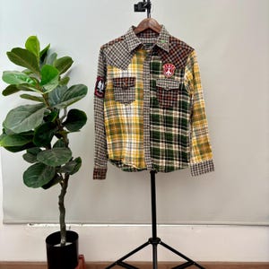 Puede incluir: Una camisa de cuadros de manga larga con diseño de patchwork, con patrones amarillos, verdes, marrones y rojos. La camisa tiene dos bolsillos en el pecho y un parche de "Peace". Se muestra en un trípode negro.