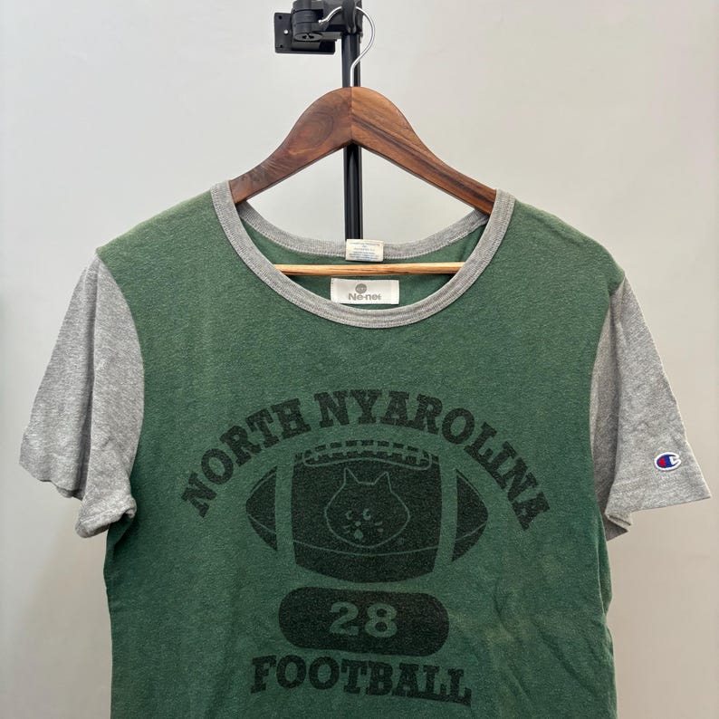 May include: A green and gray short-sleeve t-shirt with a crew neckline. The front features a black graphic of a football with a cat face inside, the text NORTH NYAROLINA, the number 28, and the word FOOTBALL. A small Champion logo is on the sleeve.
