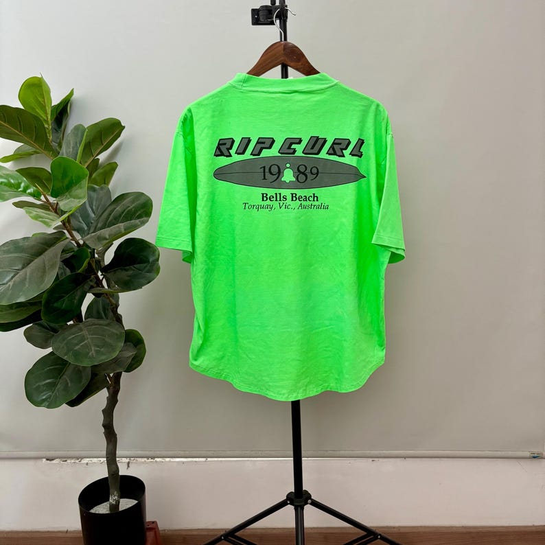 Puede incluir: Camiseta verde brillante con el texto "RIP CURL 1989 Bells Beach Torquay, Vic., Australia" impreso en la espalda. La camiseta est&aacute; colgada en una percha de madera. La camiseta es de color verde ne&oacute;n vibrante.