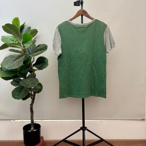 May include: A short-sleeved t-shirt featuring a solid green front panel and heather gray sleeves and neckline. The garment is displayed on a wooden hanger suspended from a black metal stand, showcasing a simple crew neck design and a straight hemline.