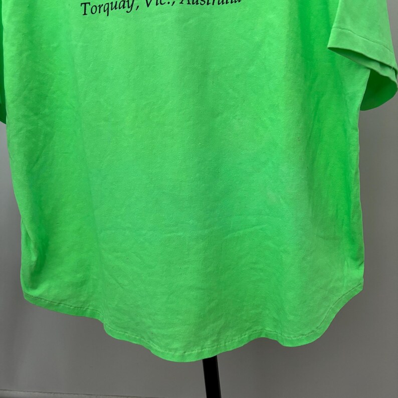 Puede incluir: Camiseta verde brillante con el texto "Torquay, Vic., Australia" en negro. La camiseta est&aacute; hecha de un material suave y tiene un dobladillo curvo. La tela tiene un aspecto ligeramente descolorido.