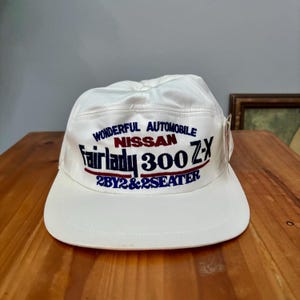 May include: A white baseball cap featuring embroidered text that reads WONDERFUL AUTOMOBILE NISSAN Fairlady 300 Z-X 2BY2&2SEATER in blue and red thread. The cap has a structured crown and a flat brim, with a small white tag attached to the side.